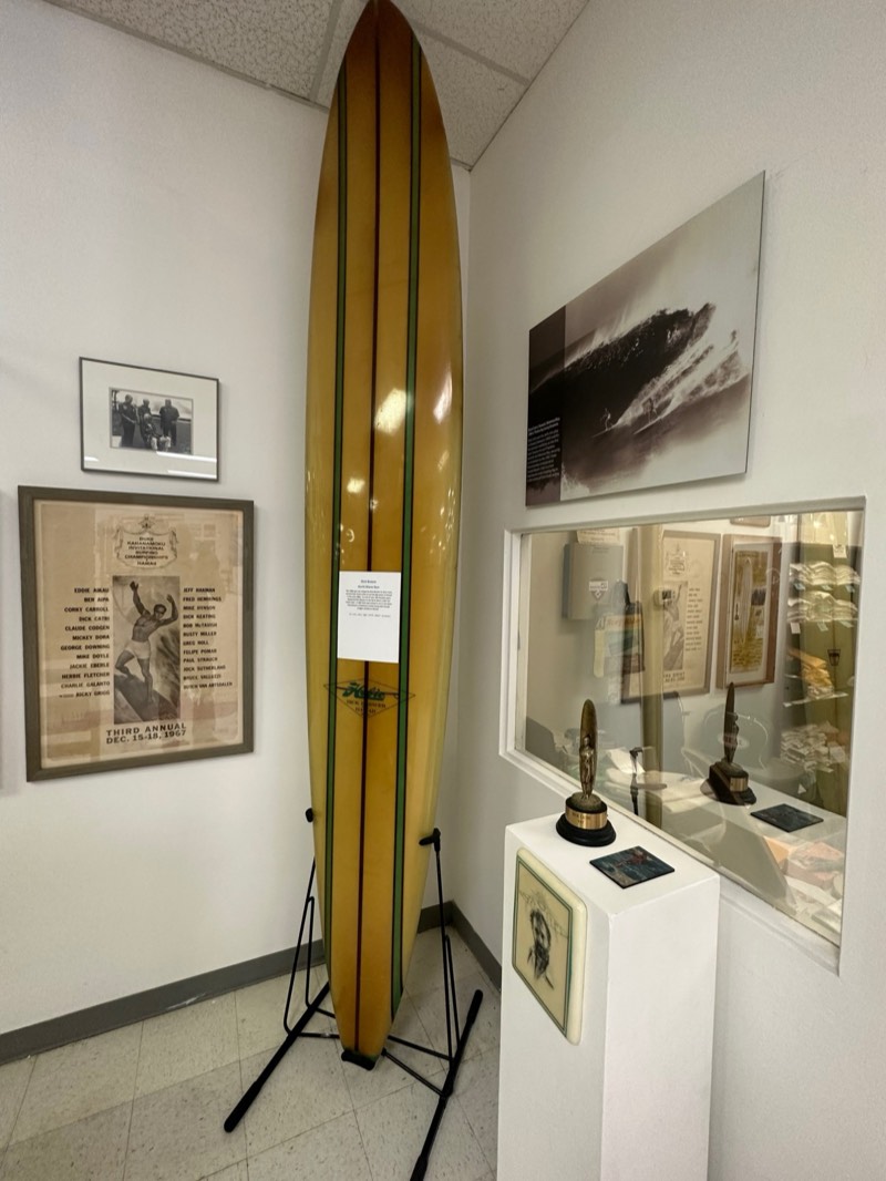 A Dick Brewer North Shore gun on display beside a 1967 Duke Kahanamoku Invitational poster and Dick Catri's trophy