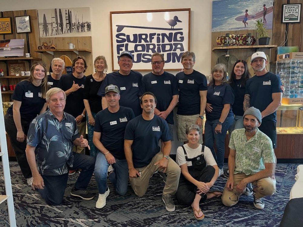 Members of the Surfing Florida Museum crew in navy museum tees, gathered in front of the sanderling sign in the gift shop